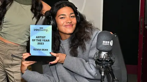 Olivia Dean holds up her BBC Introducing Award - it's a light blue glass rectangle on a black base with the words "BBC Introducing Artist of the Year 2023 Olivia Dean" written on it in black capital letters. She's sitting on an office chair behind a microphone with the Radio 1 logo on it and wearing a pair of large over-ear headphones. She smiles as she holds the award up for the camera by the base with both hands. Her long, black curly hair spills down over her shoulders and she's wearing a light grey jumper/fleece