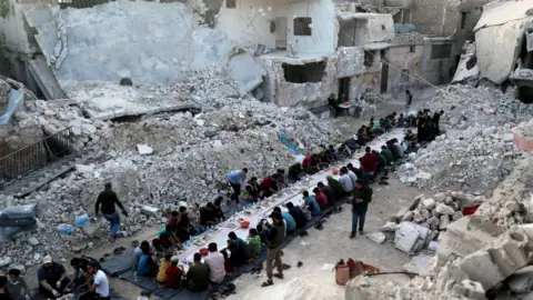 REUTERS/Khalil Ashawi People sharing a meal provided by volunteers in a bomb- damaged neighbourhood in Atarib, outside Aleppo (May 2020)