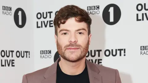 Getty Images Jordan North pictured at a BBC Radio 1 event. Jordan is a white man in his 30s with short curly brown hair and a short brown heard. He has brown eyes and looks at the camera, wearing a brown suit jacket over a black top. Behind him is white and black Radio 1 branded event hoarding