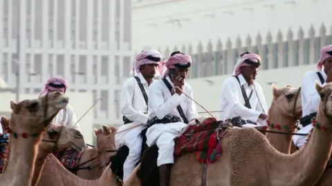 Men in the traditional Qatari clothing of white robes with red and white headscarves ride camels in front of the old parliament building in Doha