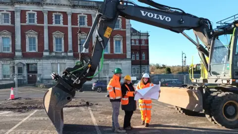 LDRS/Ted Peskett three people in construction clothing standing beneath a digger in a car park in front of a historic building