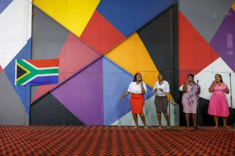 PHILL MAGAKOE/AFP People sing in front of a bright mural and the South African national flag.