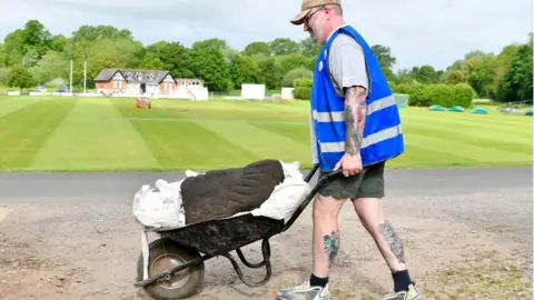 Stuart Walker Man pushes a large Roman sculpted head in a wheelbarrow