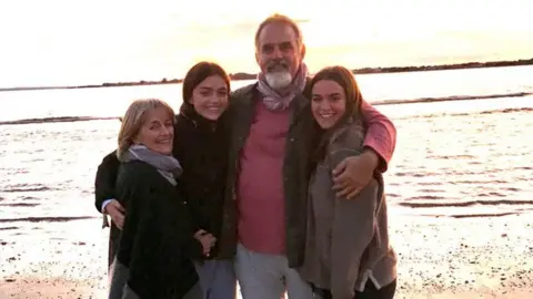 Lawrence family  A mum and dad and two young men posing on a beach smiling at the camera