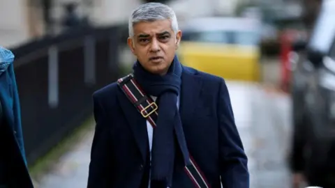 Reuters Image showing Sadiq Khan walking down a street wearing a navy blue coat and scarf and messenger bag