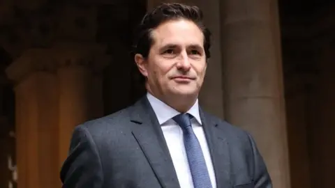 Johnny Mercer walks through a passageway wearing a suit with a blue tie