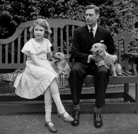 Getty Images July 1936: George, Duke of York and Princess Elizabeth sitting on a bench with their corgi dogs in the grounds of their London home, 145 Piccadilly.