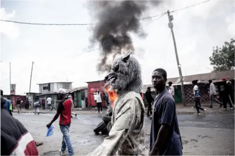 Getty Images Man walking outside next to another person who is wearing a wolf mask. There is black smoke in the background and other people who look like they are protesting or rioting.