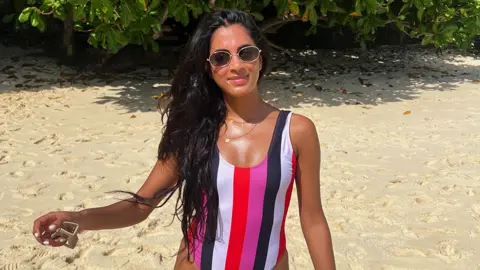Tanvi Shah Tanvi is on the beach, wearing sunglasses with the sun shining on her face. There is sand in the background with some green leaves visible from trees. Tanvi is wearing a swimsuit, which is multi coloured stripes of pink, black, white and red