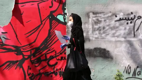 AFP A Iranian woman wearing a protective mask and gloves walks past a mask graffiti in Tehran