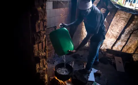 BBC/SHIRAAZ MOHAMED An unknown man pours water from a container into a pot over an open fire in preparation of cooking a meal in the derelict San Jose building in Johannesburg, South Africa