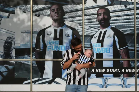 OLI SCARFF / AFP Newcastle United supporter Ryan Arrowsmith poses with three-month old son Lewis outside the club shop at St James' Park in Newcastle upon Tyne in northeast England on October 8, 2021, after the sale of the football club to a Saudi-led consortium was confirmed the previous day.