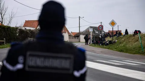 Getty Images A French gendarme standing near a group of migrants waiting for a bus in Calais after a failed crossing attempt in March 2022