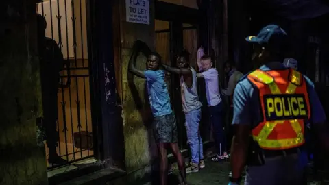 AFP Suspects are lined up against a wall as a police officer wearing gloves arrests them because they defied the lockdown order during an operation in Johannesburg, on March 27, 2020