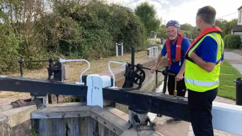Two workers from the Canal and River Trust wearing blue polo tops and life jackets stand next to the wooden top lock. Lock is made up of a wooden structure blocking the water and two large wooden bars that are used to open it on the surface. 