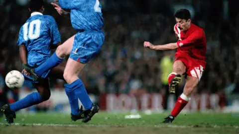 Getty Images Ian Rush, in a red Liverpool kit, shoots a ball past two defenders playing in blue kits