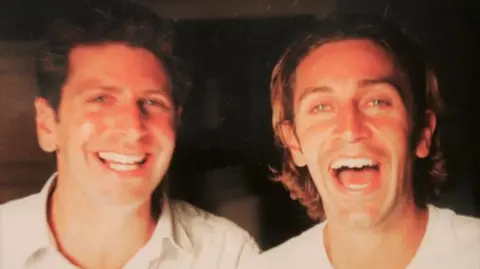 Family Handout Piers Simon on the left wearing a white collared shirt. He has dark hair, green eyes, and is smiling at the camera. His younger brother Luke is on the right, wearing a white t-shirt and laughing at the camera. He has green eyes, and shoulder-length brown hair with blonde highlights.