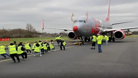 A line of people pulls on a rope that is attached to an aeroplane on a runway