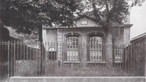 Thomas Chatterton Manuscript Project An archived image of Pile Street School in black and white. It is a modest building with two large windows at the front, with a smaller one on the right and an arched doorway on the left. There is a walkway leading up to the door and a grassy patch and trees outside the front of the building, separated from the road by spiked railings.