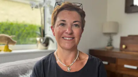 A woman with short pixie brown hair looking at the camera and smiling. She has blue eyes and a pair of glasses on her head. She has a thin pearl necklace and another necklace with a turtle pendent. She is wearing a navy blue t-shirt.