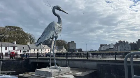 STEPH QUAYLE-JACKSON & DARREN JACKSON A giant silver heron sculpture next to a road bridge over Castletown Harbour with buildings in the background, the heron is taller than the buildings.