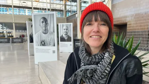 Nicola Haseler/BBC A woman in a red hat in front of a series of large black and white portrait photographs 