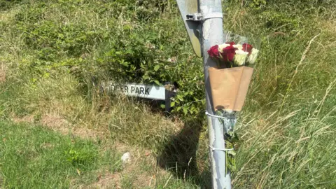 A metal post with a bouquet of flowers tied to it. In the background there is grass, and a sign which is partly covered by the long grass.