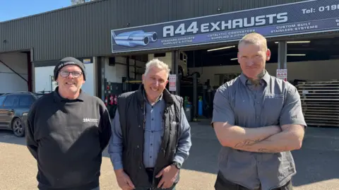 BBC Three men are looking at the camera - the man on the left is the only one wearing glasses and a black hat. The A44 Exhausts facility, including a sign with its name, is behind them.