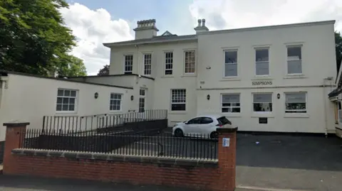 Cream-coloured two‑storey office building with “SIMPSONS” signage on the front, featuring multiple sash windows, a ramp with black railings, and a small car parked in a paved area behind a brick wall and metal fence.