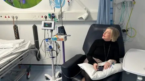 A woman with blonde hair and glasses and wearing a black top is sitting in a chair in a hospital on the right of the photo. She is looking towards equipment on the left of the image which is in front of a wall. Part of a bed is visible on the far left.