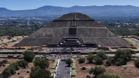 A big pyramid with mouhtains in the background, and groups of people in the foreground