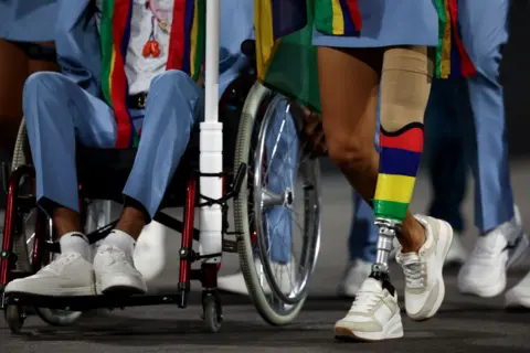 FRANCK FIFE / AFP The lower bodies of two members of the Mauritian delegation are pictured at a parade on the Place de la Concorde during the Paris 2024 Paralympic Games. One person is in a wheelchair and wearing blue trousers and white sneakers. The other has a prosthetic leg in the colors of the Mauritian national flag - yellow, green, red and blue.