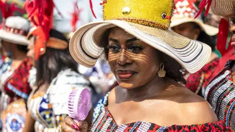 Olympia De Maismont / AFP A woman wearing a large straw hat and a hat off the shoulder holds a pink electric fan as she takes part in a festival in Calabar, Nigeria