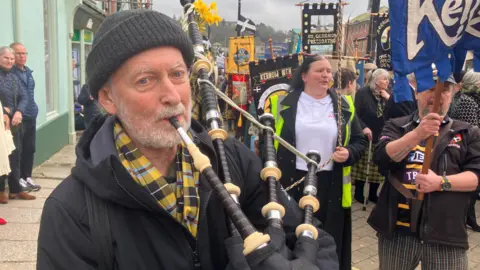 Imagen de un hombre tocando la gaita mientras participa en el desfile. Hay varias personas caminando detrás de él.
