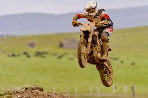 Bruce Carrington A motocross rider in a mid-air jump on bike, rider and bike are caked in mud, with a green field in the background.