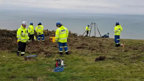 Beer Coastguard Rescue Team Seven helmeted rope technicians in hi-vis jackets stand near an array of climbing ropes draped over a cliff edge