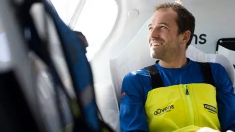 A bearded man in a blue and yellow sailing outfit in the cockpit of a yacht.