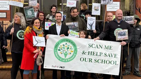 Martin Heath/BBC A group of adults and children posing outside the brick-built offices of Hertfordshire County Council in Stevenage. They are holding signs and a large white banner.