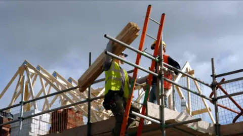 Two construction workers in high-visibility clothing and hard hats are on scaffolding at a building site. One is carrying wooden planks on their shoulder while the other stands on the platform beside an orange ladder, with roof trusses and stacked bricks visible in the background under a cloudy sky.