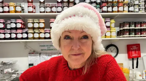 A woman with blonde hair looking into the camera, wearing a red woollen jumper and a pink Santa hat. Behind her are rows of jams and preserves; she is standing behind her counter in Wellington Market.