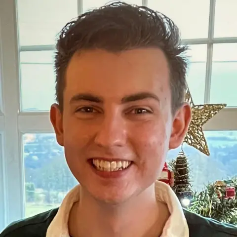 Tracy Scott Ethan, who has short brown hair, looking directly at the camera and smiling. There is a window and a Christmas tree behind him.