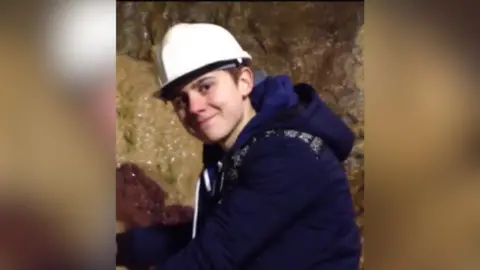 Lisa Bowerman A teenage boy, wearing a white helmet and navy hoodie, smiles inside a cave.