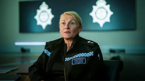 Yeti Television/BBC Cymru Wales Emma White is a white woman in middle age. She has dyed blonde hair pulled back into a low bun or ponytail. She wears a black zip-up jacket with two strips of blue and white check pattern across the best and arms with a badge reading "Heddlu Police" underneath the right one as the camera sees it. She is lightly made up. Behind her slightly out of focus is an office with a large screen on the wall, with two silver police crests displayed on it. 