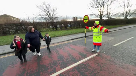 Maureen Seaton standing in the road with her lollipop stop sign held out. A mother and her children, a boy and a girl, are crossing the road outside of Coatham Primary School.