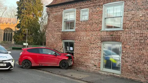 Mark Nundy A red car has crashed into the window and wall of an old pub building. A police car can be seen to the side of the crash. 