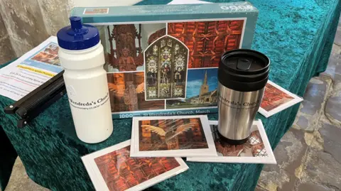 John Devine/BBC A selection of merchandise on a trestle table inside the church, including a 1000-piece jigsaw puzzle of St Wendreda's, and some water bottles and postcards bearing its image.