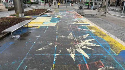 The pavement has been painted with blue, red, yellow and white star-shaped and circular patterns. Black patches are showing through in several places. People are walking in the distance.