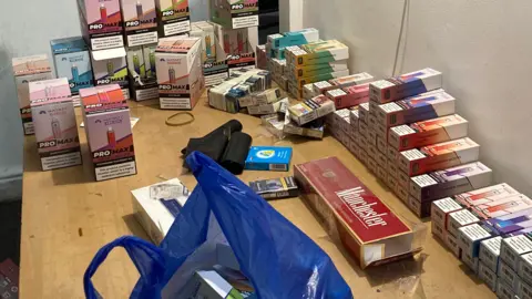 A wooden table on which are sitting many cigarette packets and cartons as well as boxes of disposable vapes.