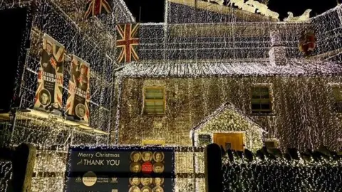 Taxi Charity for Military Veterans Richard Iles' house completely covered in white string lights. A sign attached to a gate says Merry Christmas and a thank you message to veterans. Union Jack flags are also hung from his home.