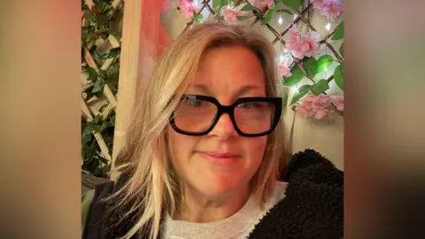 Claire Unwin A close-up of Claire in front of a trellis with pink flowers on it. She is slimmer than in the first image, with styled blonde, shoulder-length hair and black glasses with large, bold frames.